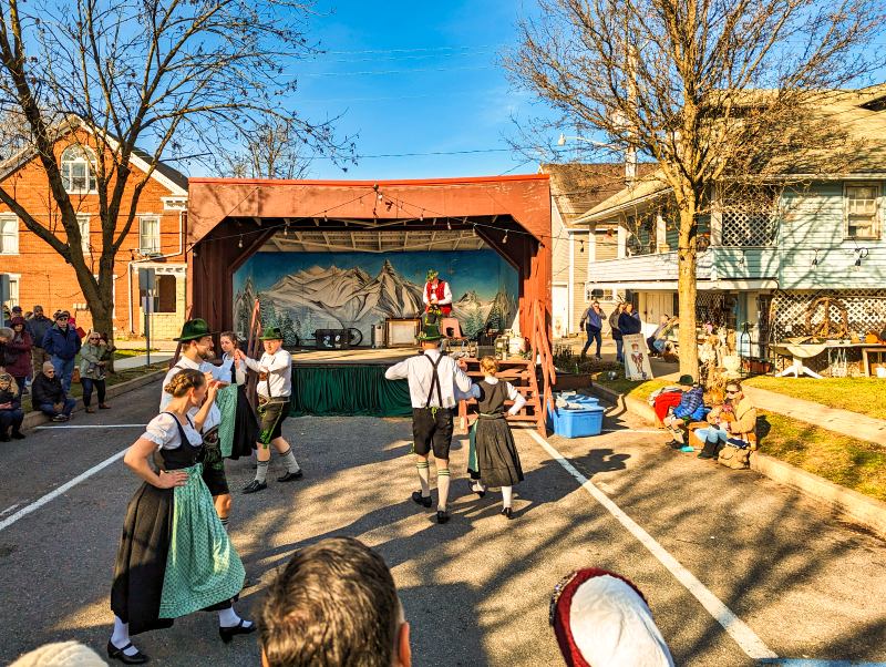 Performers in traditional German attire dancing in front of a stage with a scenic backdrop, while onlookers watch and enjoy at the Mifflinburg Christkindl Market in Mifflinburg, Pennsylvania, with residential buildings in the background under a clear sky.