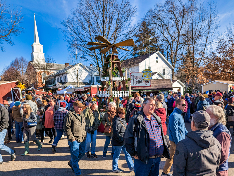 Crowded scene of visitors enjoying the Mifflinburg Christkindl Market in Mifflinburg, Pennsylvania, with a decorative windmill, "Happy Holidays" sign, white church steeple in the background, and people mingling under a clear blue sky.
