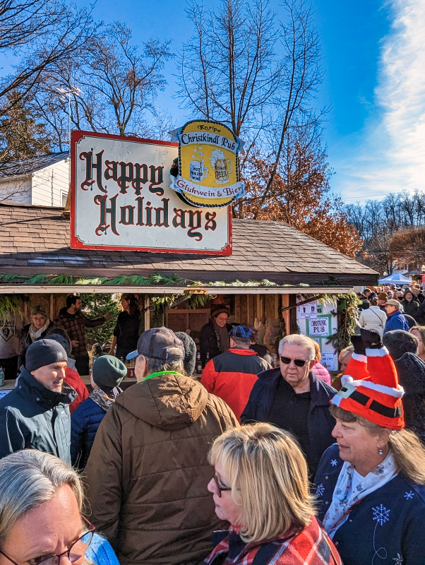 Crowd of visitors at the Mifflinburg Christkindl Market under a 'Happy Holidays' sign, with a direction sign to the 'Christkindl Pub' serving 'Glühwein & Bier' in the background, indicating a festive and bustling holiday atmosphere.