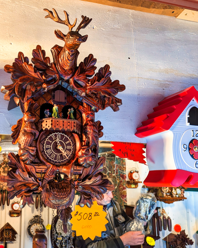 Elaborate wooden cuckoo clock with deer head adornment and intricate carvings, priced at $508.30, displayed at a stall at the Mifflinburg Christkindl Market, with other clocks in the background and a shopper perusing items.