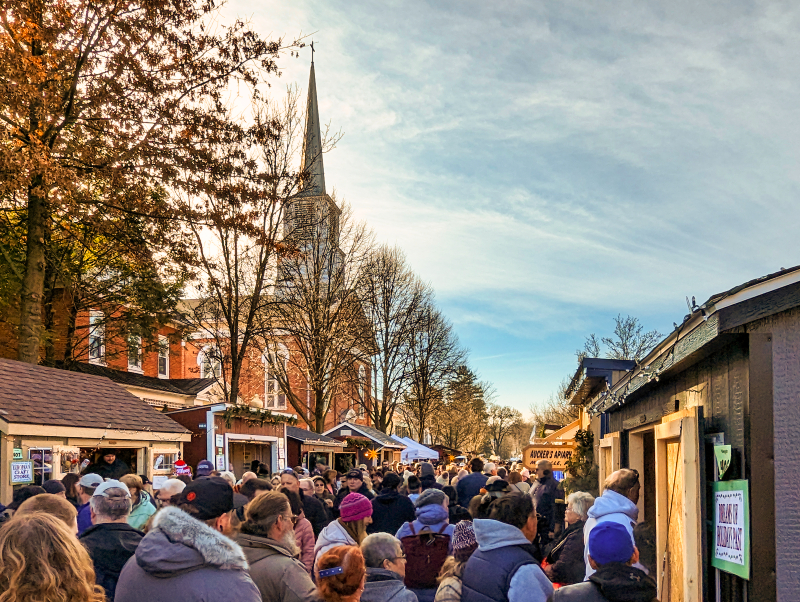Crowded pathway at the Mifflinburg Christkindl Market with visitors browsing stalls, a tall church steeple in the background, and autumn trees lining the scene under a partly cloudy sky.