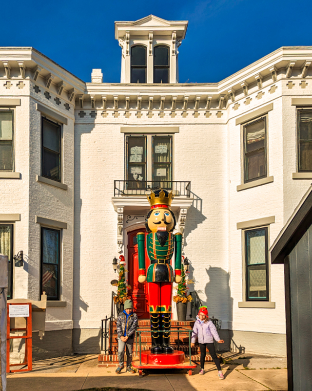Large festive nutcracker statue positioned in front of a white historic building with two individuals posing beside it, under a clear blue sky at the Mifflinburg Christkindl Market in Mifflinburg, Pennsylvania.