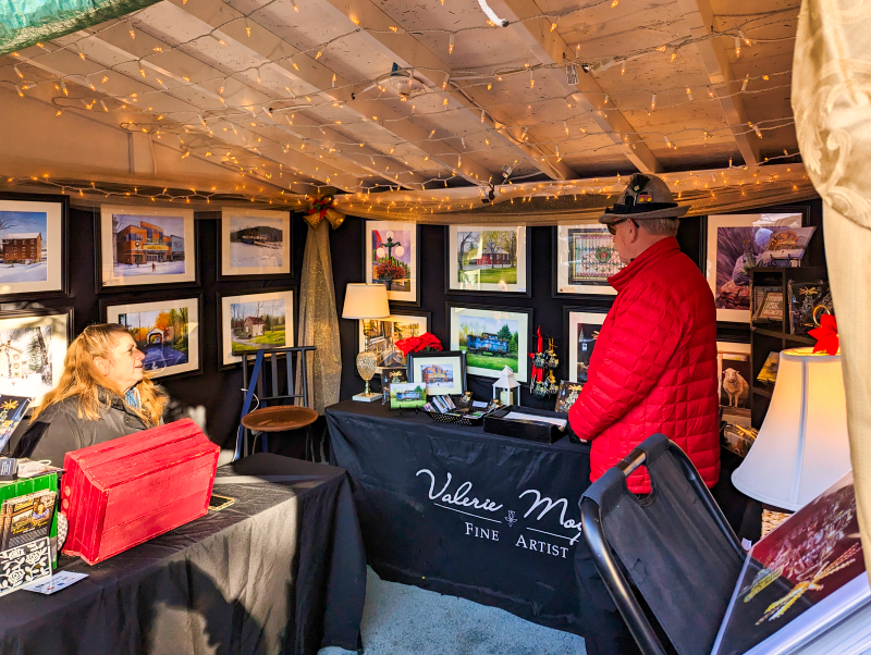 An artist's booth illuminated with fairy lights at the Mifflinburg Christkindl Market in Mifflinburg, Pennsylvania, showcasing framed artworks on the wall, with a seated female artist and a male visitor wearing a red jacket observing the pieces.