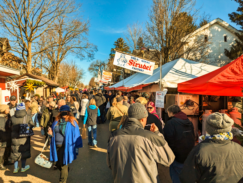 Crowds of visitors exploring the lively Mifflinburg Christkindl Market, with a prominent sign for 'The Original Strudel Factory' amidst stalls, under a clear blue sky in Mifflinburg, Pennsylvania.