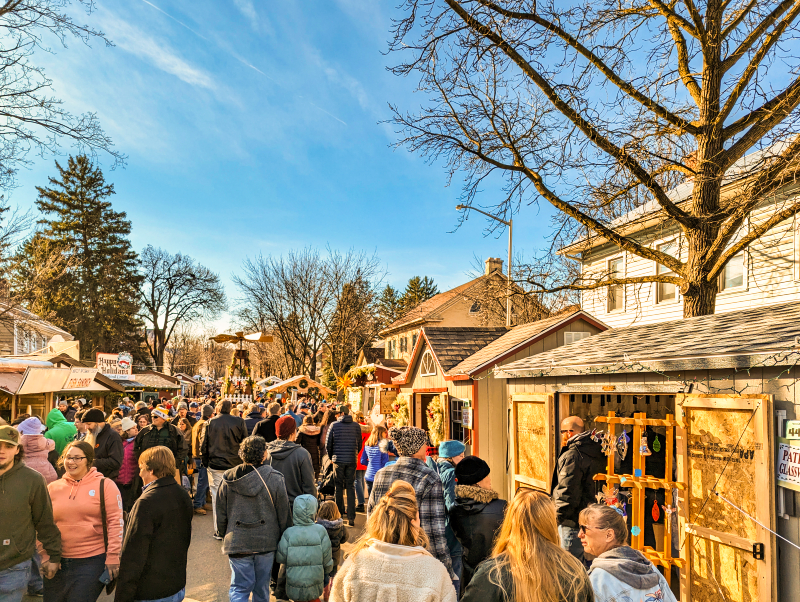 Crowded street at Mifflinburg Christkindl Market with visitors exploring wooden stalls, under a clear blue sky with historic homes in the background and leafless trees overhead in Mifflinburg, Pennsylvania