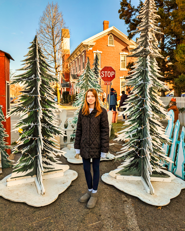 Young girl standing between two decorative wooden Christmas trees with a historic red-bricked building in the background, at the Mifflinburg Christkindl Market during daytime.
