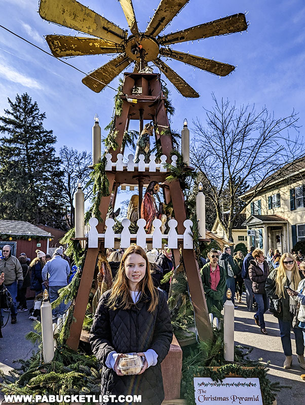 Young girl standing in front of a large Christmas pyramid adorned with figurines and candles, with a rotating windmill top, surrounded by visitors at the Mifflinburg Christkindl Market under a blue sky.