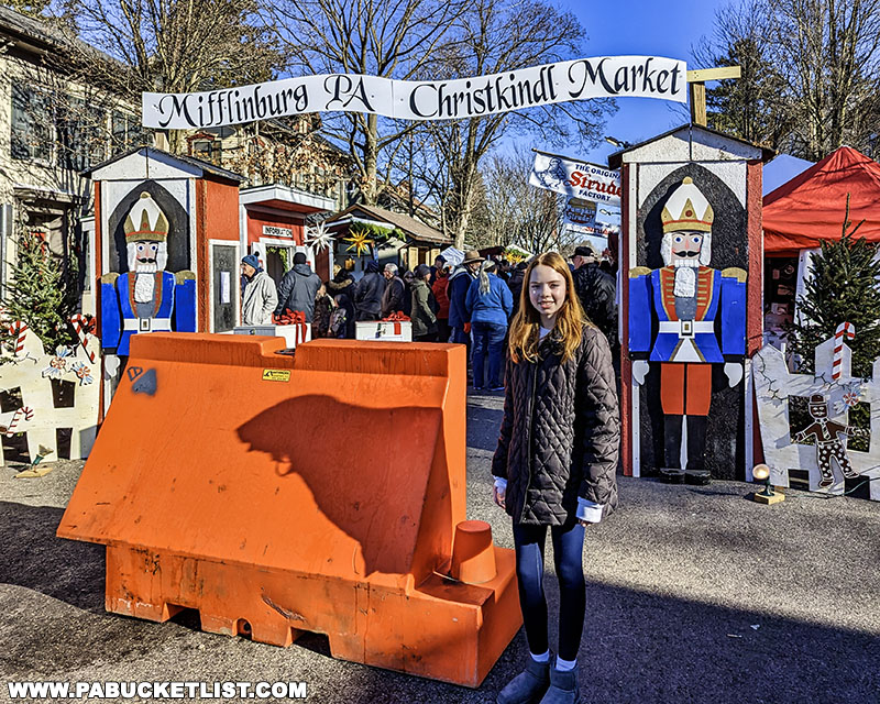 Young girl standing beside entrance to the Mifflinburg Christkindl Market, with wooden nutcracker decorations, market stalls, and visitors in the background under a clear sky.