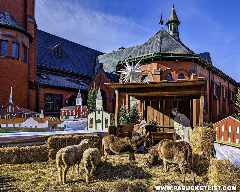 Outdoor nativity scene at the Mifflinburg Christkindl Market, featuring live sheep and donkeys in front of a red brick church, with wooden cutouts depicting Bethlehem buildings.