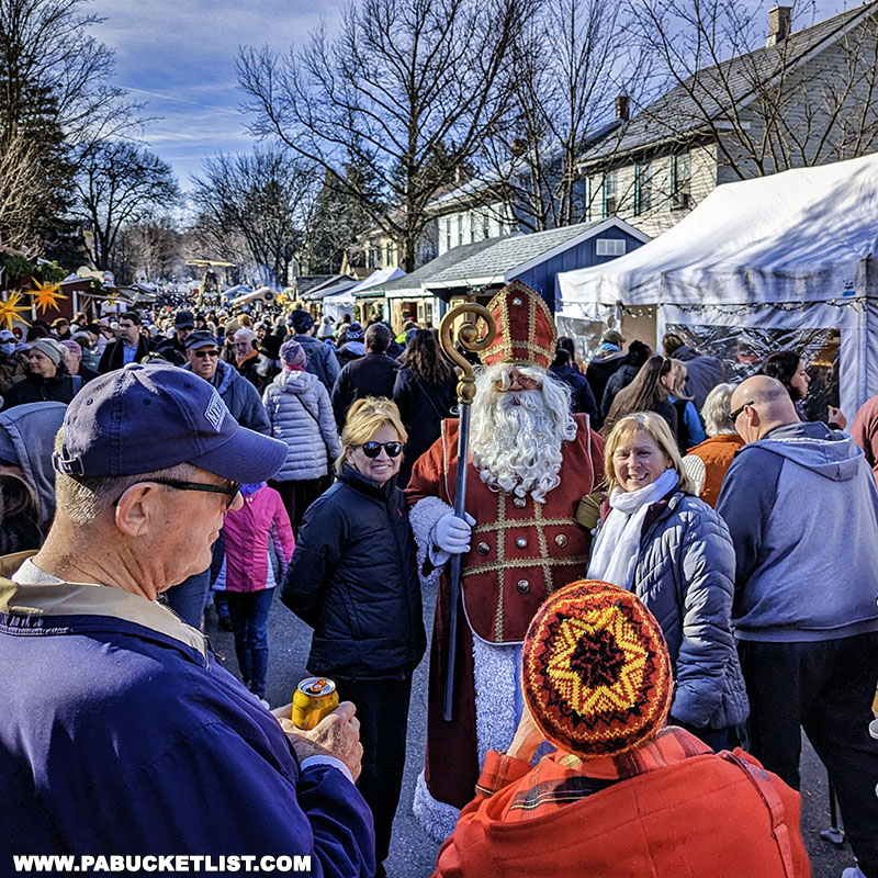 Crowded scene at the Mifflinburg Christkindl Market with a figure dressed as Saint Nicholas holding a staff, while attendees mingle and browse stalls under a clear blue sky.
