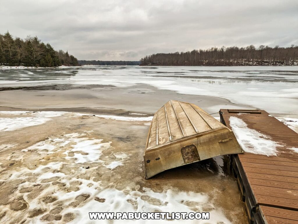 A winter scene at Lake Jean near the Mountain Springs - Cherry Run loop in Ricketts Glen State Park. An overturned aluminum rowboat rests on a snowy dock, with the icy surface of the lake stretching into the distance. The surrounding landscape is quiet and subdued, with leafless trees lining the far shore under a pale, overcast sky. Patches of snow and ice on the ground near the dock hint at the cold, still atmosphere of the season.