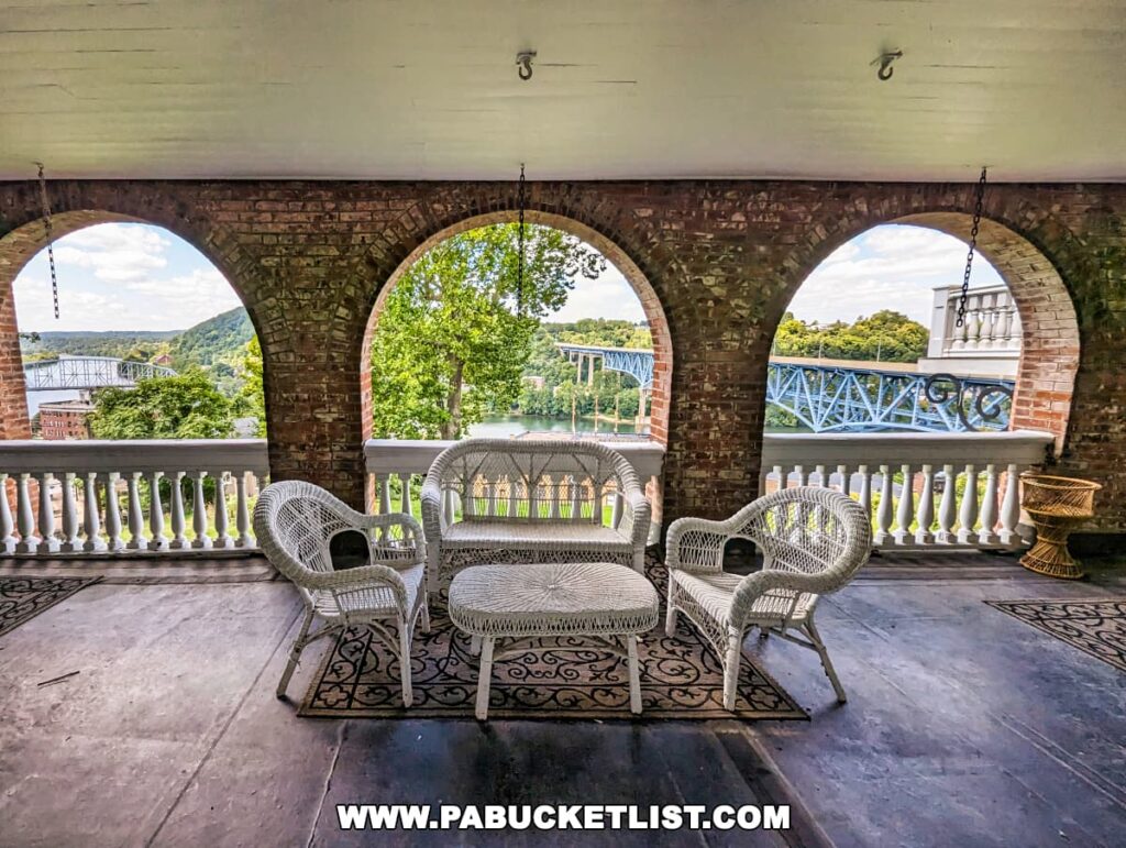 Elegant porch with white wicker furniture and patterned rugs at Nemacolin Castle, showcasing a scenic view of the landscape and a bridge, framed by arched brick openings.
