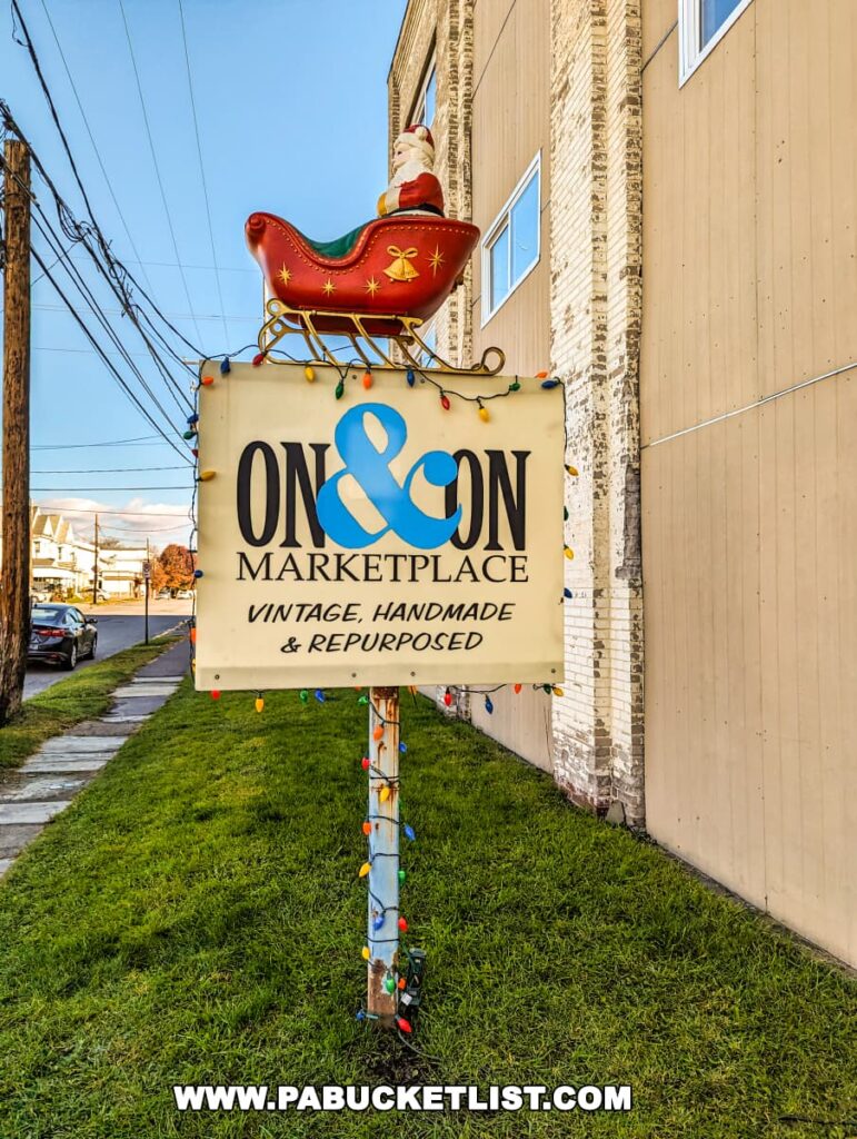 A colorful sign for the 'ON&ON MARKETPLACE' with the words 'VINTAGE, HANDMADE & REPURPOSED' below. The sign, adorned with string lights, is mounted on a pole and topped with a whimsical red sleigh and Santa figure. The sign is positioned against the side of a tan building with a clear blue sky above, and it stands over a well-kept grassy area.