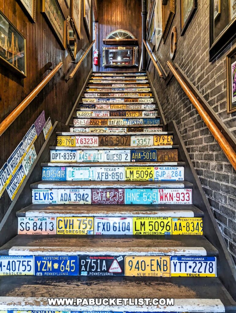 An interior staircase at the On and On Vintage Marketplace in Scranton, Pennsylvania, creatively decorated with a variety of colorful vintage license plates from different states on each step. The walls are adorned with framed pictures, and the warm lighting accentuates the wooden textures of the stairway and wall paneling.