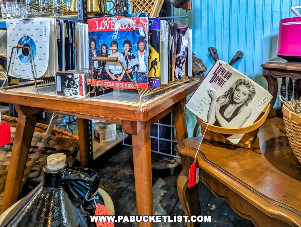 A vintage record display at the On and On Vintage Marketplace featuring classic vinyl albums, including 'Love Boys' and 'Taylor Dayne', showcased on a wooden table. The records are propped up in wooden stands for easy browsing, with other albums stored upright in the background. A large black ceramic jug with a red price tag is also visible in the foreground. The blue-paneled wall and warm wood furniture add to the nostalgic ambiance of the shop.