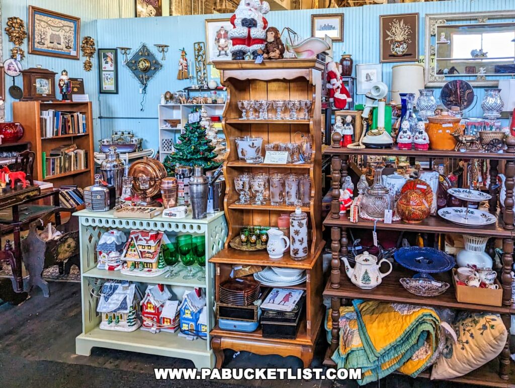 A richly decorated space within the On and On Vintage Marketplace, filled with an assortment of items. Shelves and tables display everything from holiday-themed decorations and a miniature Christmas tree to vintage tableware, glassware, and framed artwork. The area is a treasure trove of collectibles with a quaint, festive ambiance, featuring wooden furniture and a blue corrugated wall. A variety of lamps and a Santa figure add to the eclectic mix.