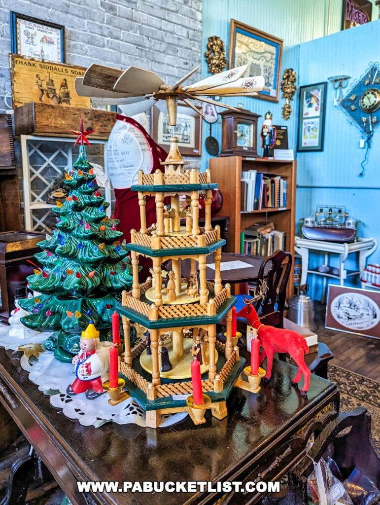 A festive display at the On and On Vintage Marketplace featuring a traditional German Christmas pyramid with spinning fan blades powered by candle heat, set upon a wooden table. Beside it is a ceramic Christmas tree and a vintage toy clown. In the background, various antique items and framed pictures contribute to the shop's nostalgic atmosphere. The blue wall panels and vintage advertising signs add to the charm.