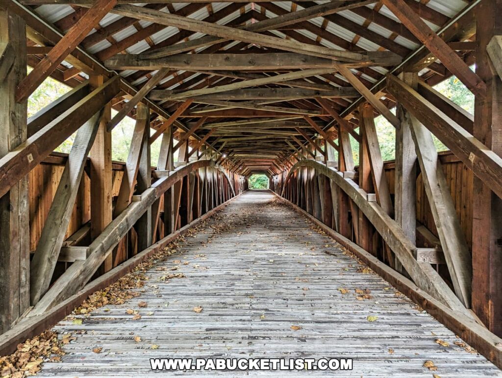 Interior view of the Pomeroy-Academia Covered Bridge in Juniata County, Pennsylvania, highlighting the intricate wooden Burr arch truss design. The worn wooden planks of the bridge floor are scattered with fallen leaves, suggesting the autumn season, and the perspective leads the viewer's eye through the length of the bridge to the light at the far end.