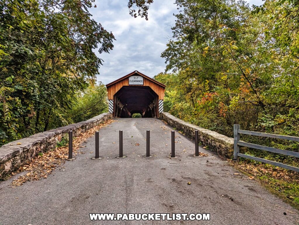 The entrance to the Pomeroy-Academia Covered Bridge in Juniata County, Pennsylvania, viewed from the road leading up to it. The bridge is marked with a '11.7 FT CLEARANCE' sign and is closed to vehicular traffic, as indicated by the series of black bollards on the road. Autumn leaves dot the ground and lush green trees with hints of fall colors frame the scene.