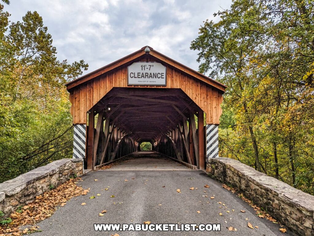 Frontal view of the Pomeroy-Academia Covered Bridge in Juniata County, Pennsylvania, with '11-7" CLEARANCE' sign prominently displayed above the entrance. The bridge features a traditional wooden design with a stone foundation on either side. Autumn leaves are scattered on the road leading to the bridge, and the surrounding foliage is starting to show fall colors.
