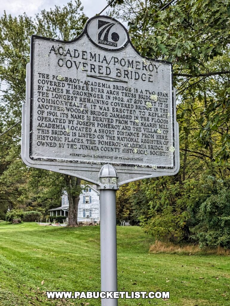 Close-up of the historical marker for the Academia/Pomeroy Covered Bridge in Juniata County, Pennsylvania. The sign, marked with the PennDOT logo, provides historical details about the bridge, mentioning its construction with a Burr arch truss design in 1902 and its status as the longest remaining covered bridge in the state. Lush green trees surround the sign with a traditional two-story house visible in the background.