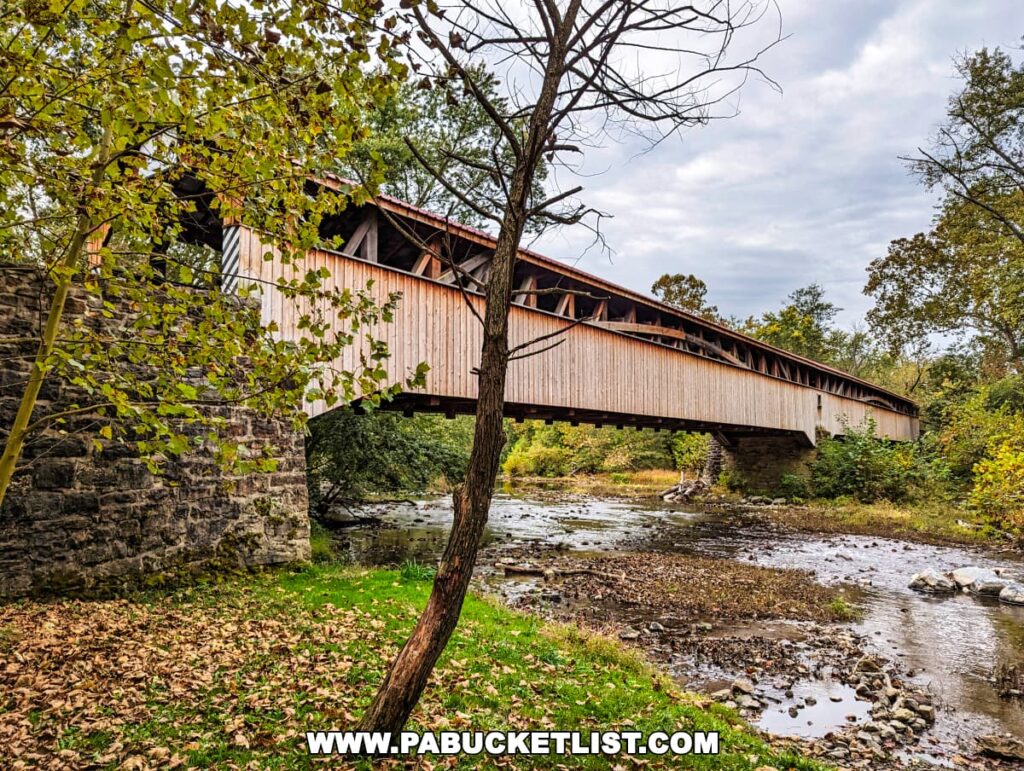 Side view of the Pomeroy-Academia Covered Bridge near Port Royal in Juniata County, Pennsylvania, spanning a shallow creek. The historic wooden bridge rests on stone foundations and is surrounded by a landscape with early fall foliage and fallen leaves. Overcast skies and the serene creek contribute to the peacefulness of the scene.
