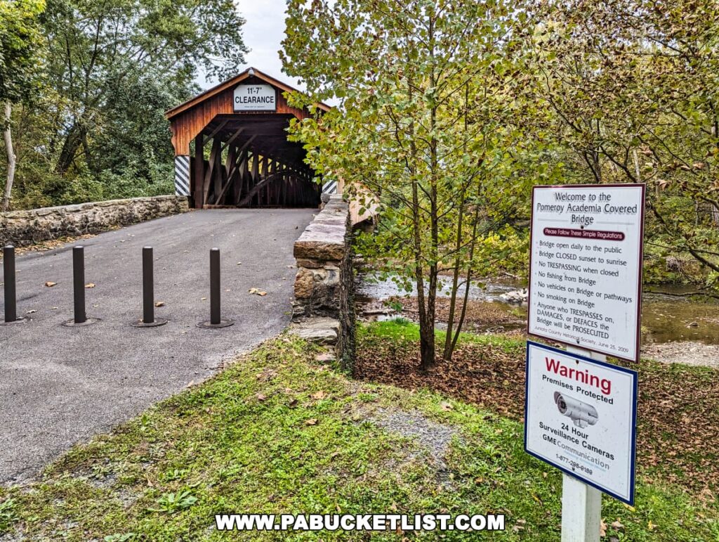 The entrance to the Pomeroy-Academia Covered Bridge in Juniata County, Pennsylvania, with a series of black bollards in the foreground indicating that the bridge is closed to vehicular traffic. Next to the bridge is a sign with rules for visitors, including no trespassing when closed, no fishing from the bridge, and no smoking on the bridge or pathways, reinforcing its protected status. Another sign warns of 24-hour surveillance. The surrounding area is lush with trees showing early fall colors.