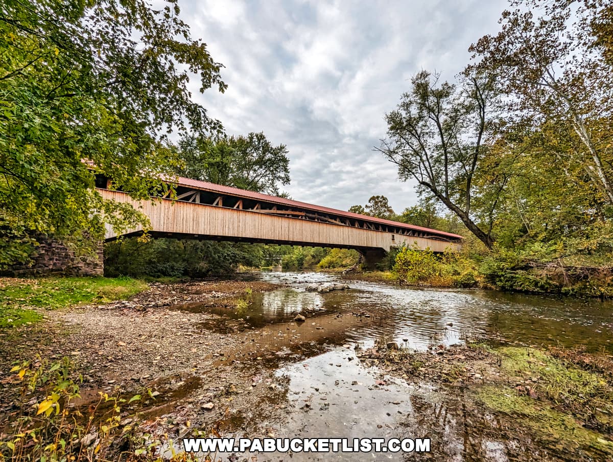 Side angle view of the Pomeroy-Academia Covered Bridge in Juniata County, Pennsylvania, extending over a creek with running water. The bridge's wooden structure is supported by stone abutments and framed by trees with leaves beginning to change for autumn. The cloudy sky adds to the serene and rustic atmosphere of the setting.