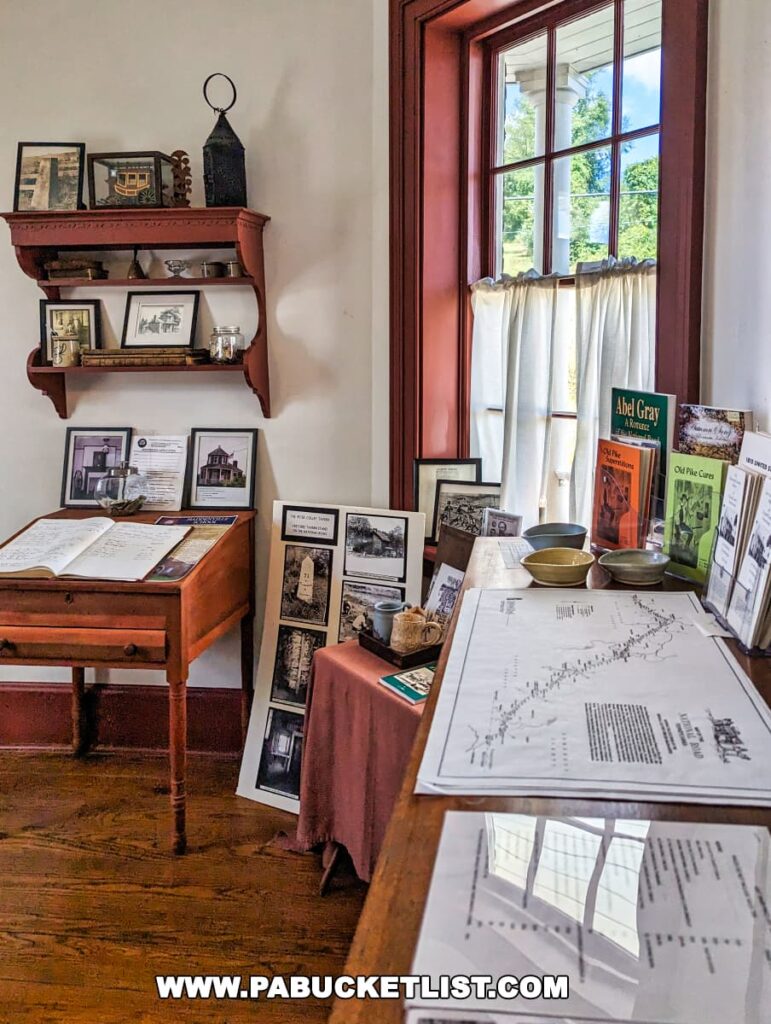 A cozy interior corner of the Searights Tollhouse in Fayette County, Pennsylvania, featuring a historical display. A wooden writing desk is adorned with open ledger books, while a shelf above holds various framed images and a lantern. To the right, a tall window with red frames and white curtains allows natural light to flood the room. On a table draped with a red cloth, more framed pictures, books, and artifacts are presented, with large maps spread out for viewing.