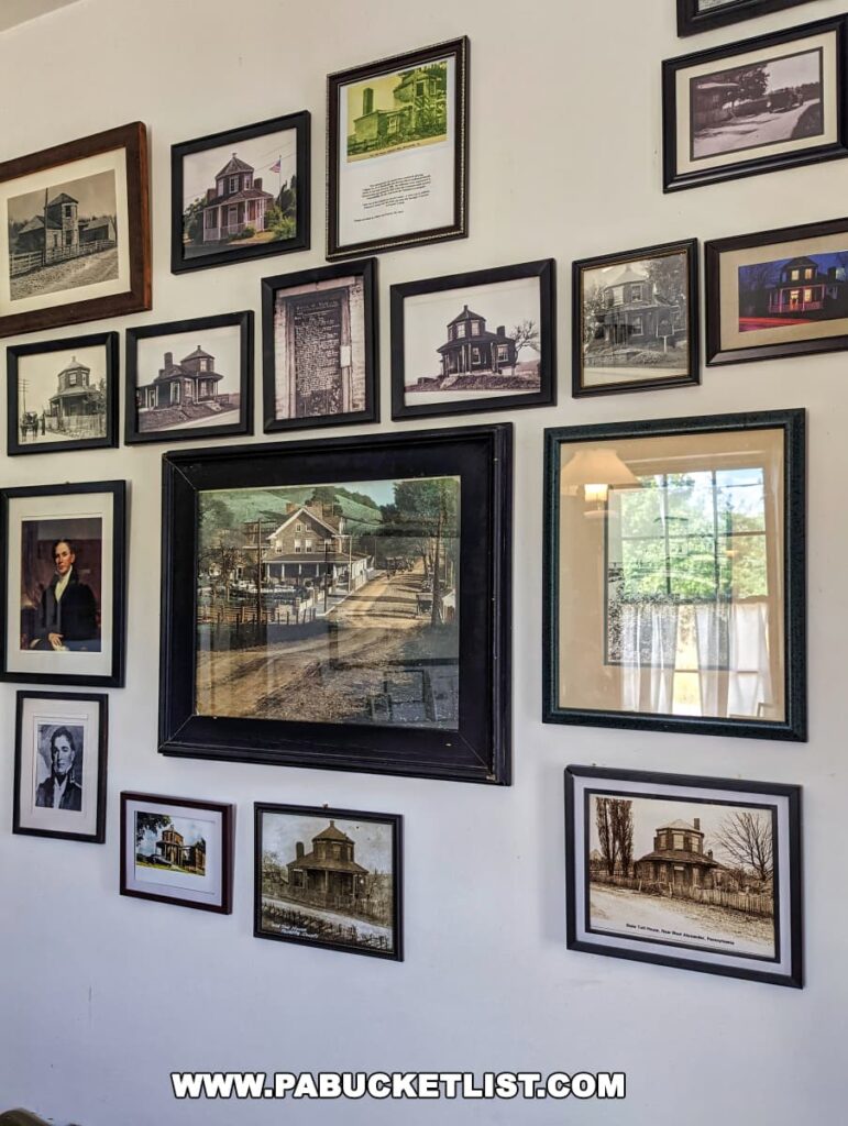 Interior wall of the Searights Tollhouse in Fayette County, Pennsylvania, adorned with a collage of framed historical photographs and portraits. Various images depict the tollhouse and other historic buildings in black and white and sepia tones, showcasing different eras and architectural details. A central, larger colored photograph draws the eye, featuring a vintage street scene. Natural light from a window is reflected in some frames, adding brightness to the display.