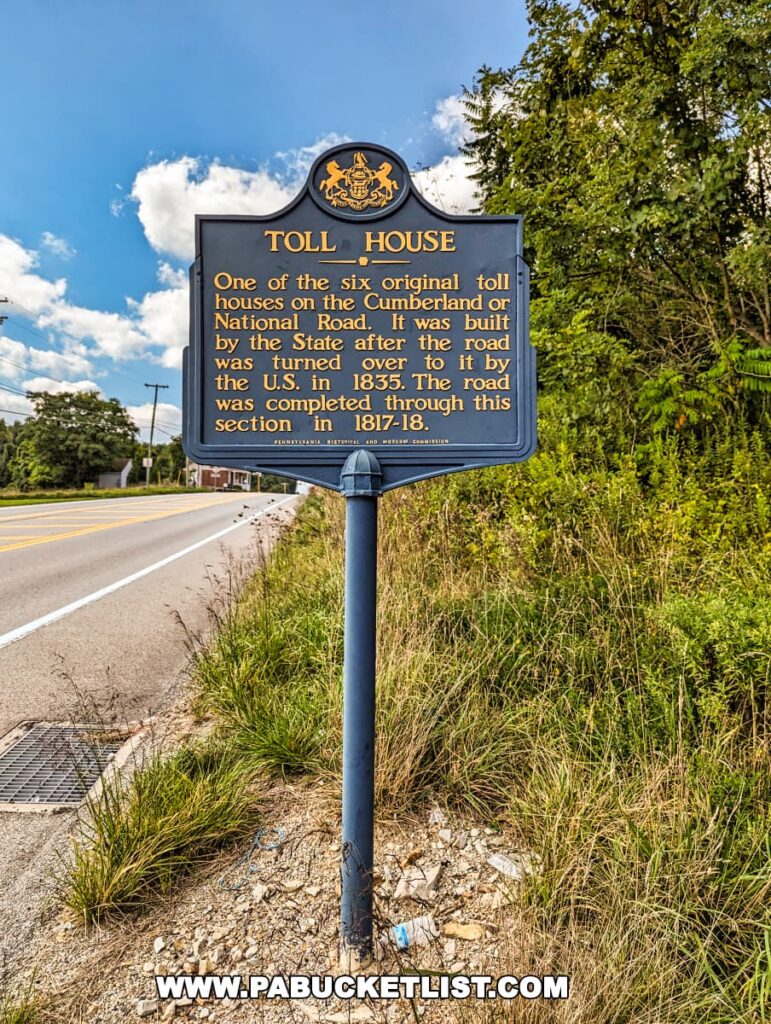 An informative historical marker sign for the Searights Tollhouse along the National Road in Fayette County, Pennsylvania. The sign has a dark background with white and yellow text, detailing the history of the tollhouse as one of the six original toll houses on the Cumberland Road, built by the State in 1835. It stands beside a road, with lush greenery in the background and a clear blue sky with clouds above.