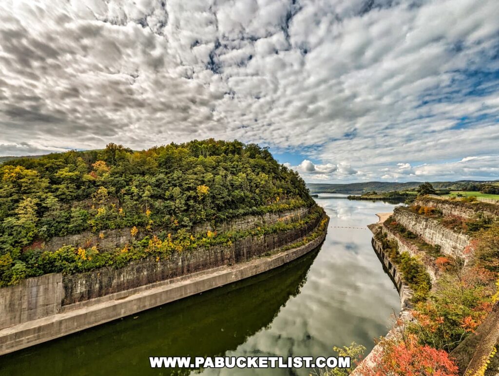 A captivating view from the Tioga-Hammond Lakes Overlook in Tioga County, Pennsylvania, featuring the connecting channel between two lakes, bordered by steep cliffs adorned with trees displaying early autumn colors, and a dramatic sky reflecting on the calm water surface.