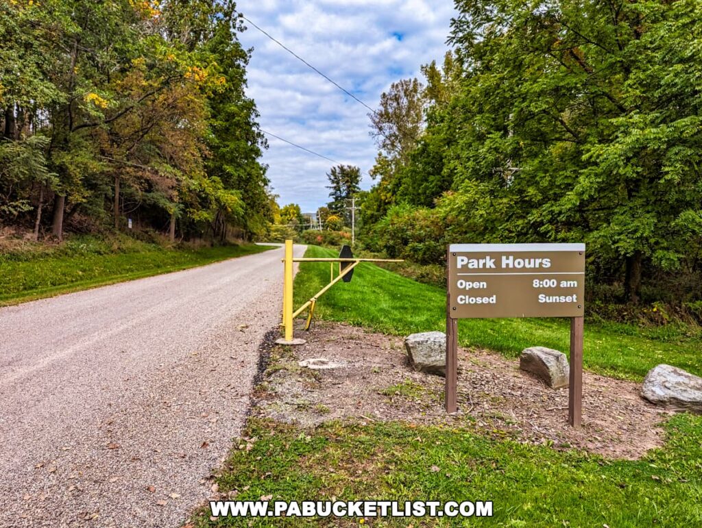 The entrance to the Tioga-Hammond Lakes Overlook in Tioga County, Pennsylvania, with a 'Park Hours' sign indicating open hours from 8:00 am to sunset beside a gravel access road, flanked by lush green trees and a closed yellow gate across the road.