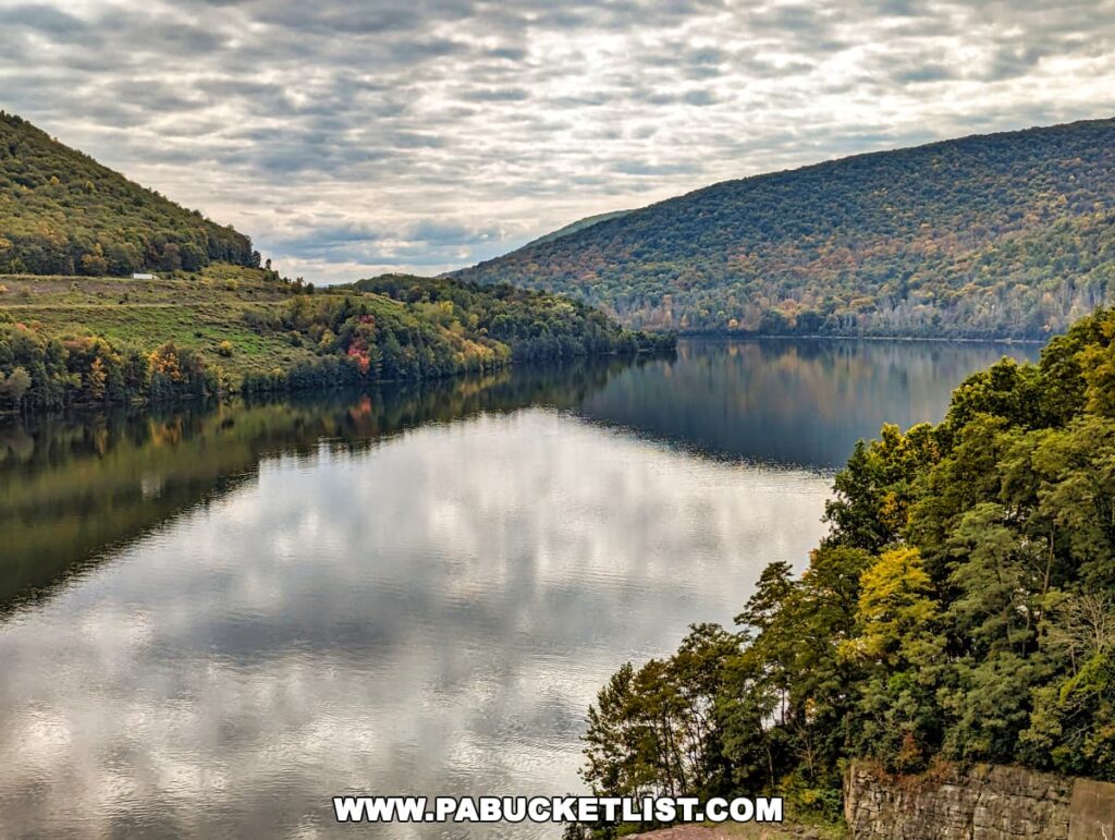 A tranquil view over Tioga Lake from the Tioga-Hammond Overlook in Tioga County, Pennsylvania, with the smooth lake waters mirroring the overcast sky and surrounding foliage-covered hills that are speckled with the warm colors of fall.