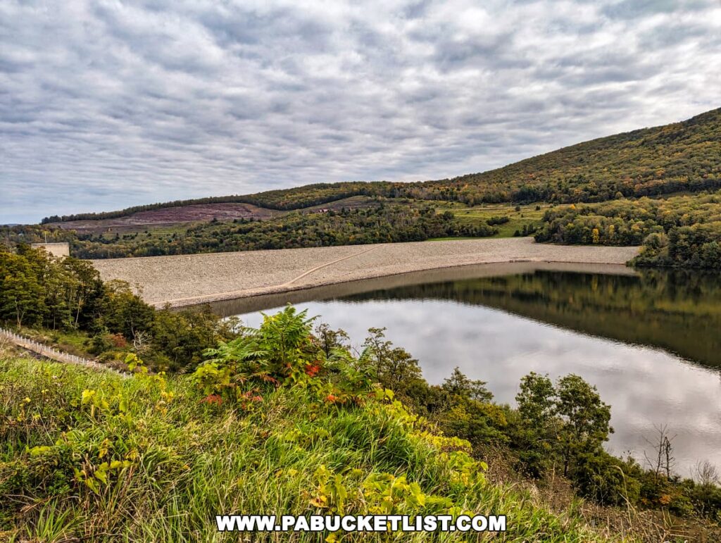 View of the dam at Tioga Lake from the Tioga-Hammond Overlook in Tioga County, Pennsylvania, showcasing the dam's large rock-filled structure reflecting on the calm lake surface, with surrounding hills displaying the changing colors of early autumn under a cloudy sky.