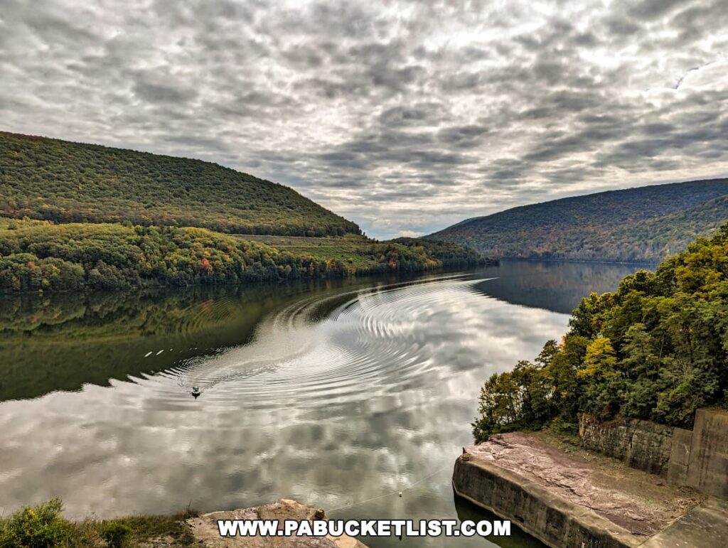 Scenic view from Tioga-Hammond Lakes Overlook in Tioga County, Pennsylvania, displaying a calm reservoir with a single boat creating ripples on the water, surrounded by lush hills reflecting in the water, under a sky partially covered with fluffy clouds.