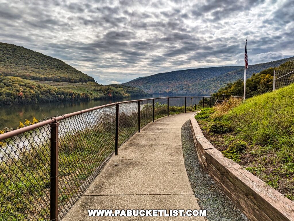 A concrete walkway leading to the viewing area at the Tioga-Hammond Lakes Overlook in Tioga County, Pennsylvania, with a chain-link fence on one side and a hill with green grass on the other. The American flag waves in the distance, with a serene river and forested hills under a cloudy sky in the background.