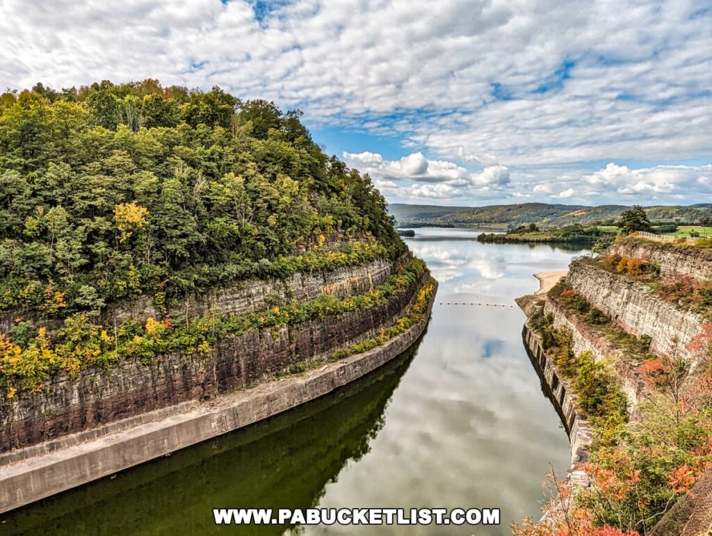 A striking view from the Tioga-Hammond Lakes Overlook in Tioga County, Pennsylvania, showcasing a water channel cutting through steep cliffs covered with a mix of green and autumn-colored trees, reflecting the sky in the still water, with expansive views of the landscape under a patterned cloud cover.