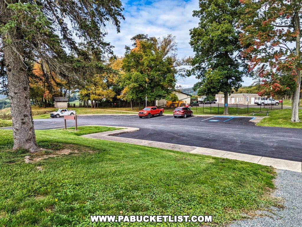 A parking area at Tioga-Hammond Lakes Overlook in Tioga County, Pennsylvania, showing several parked cars, freshly painted parking lines, handicap-accessible spaces, and a background of trees displaying early fall colors. A grassy area with mature trees frames the foreground.