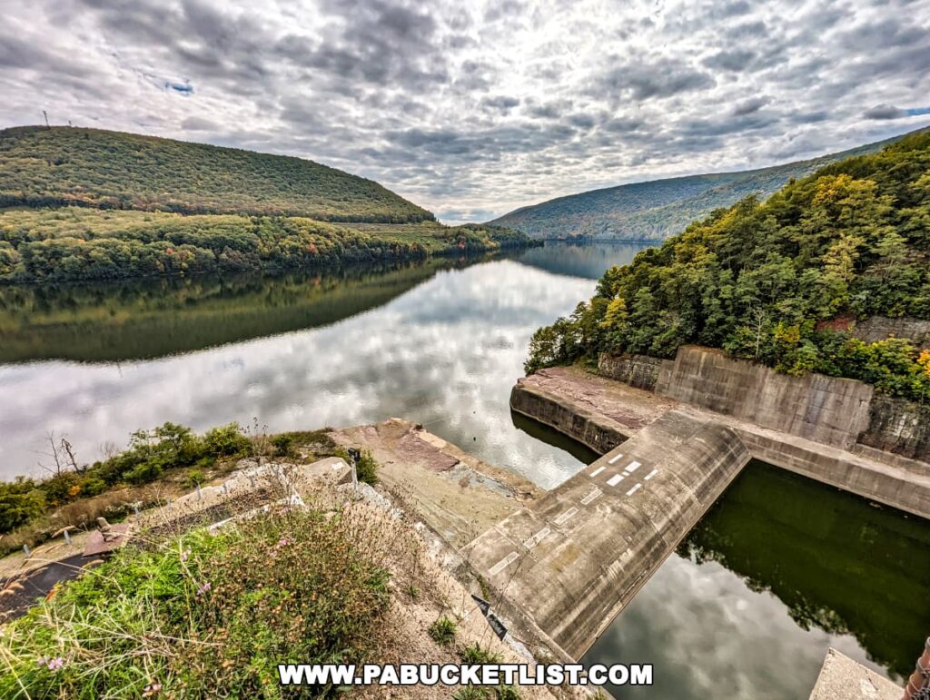 View from the Tioga-Hammond Lakes Overlook in Tioga County, Pennsylvania, featuring a serene Tioga Lake with a reflection of the overcast sky, flanked by lush, green hillsides with hints of fall foliage, and a concrete structure at the water's edge.