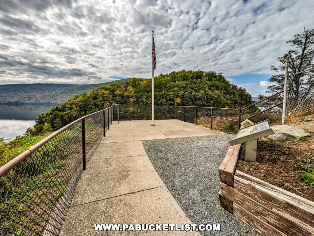 The walkway leading to the viewing platform at the Tioga-Hammond Lakes Overlook in Tioga County, Pennsylvania, with a chain-link fence on one side, an American flag flying high, and an informative plaque mounted on a stone pedestal. The overlook provides a view of the lake and forested hills under a dramatic, cloud-filled sky.