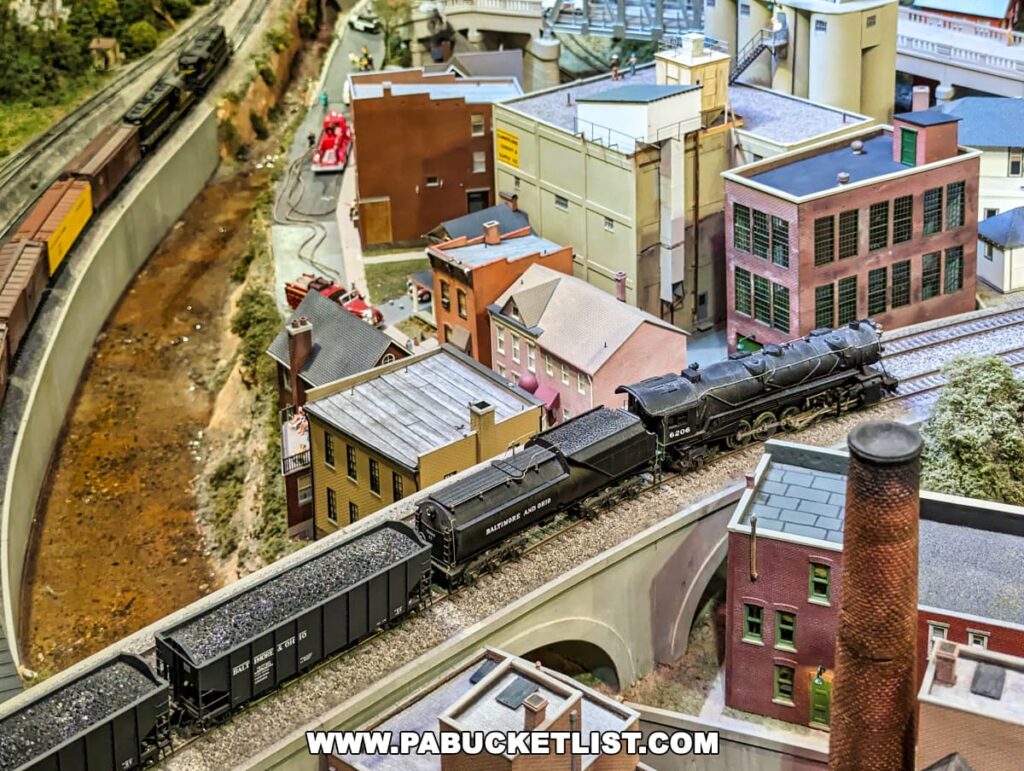 A close-up of a meticulously detailed model train setup at the Western PA Model Railroad Museum. The scene features a miniature B&O (Baltimore and Ohio) locomotive with a black engine and coal car pulling a line of freight cars through a scaled-down industrial town. The town includes realistic buildings such as brick houses, industrial facilities, and a factory with a tall smokestack. The layout also features a riverbed parallel to the tracks and tiny figurines that bring the miniature world to life, all showcasing the museum's commitment to capturing the essence of a bygone era in railroad history.