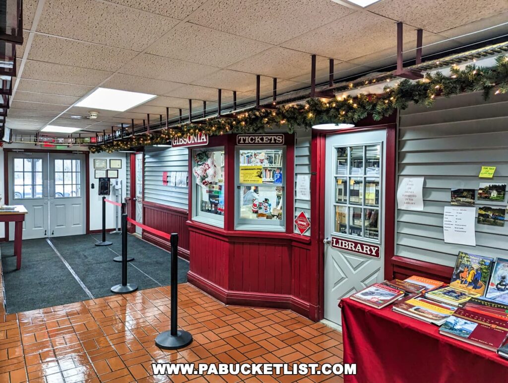 Lobby area of the Western PA Model Railroad Museum in Gibsonia, featuring a ticket window with 'GIBSONIA' signage, festive garland decorations, a library door to the right, various train-themed books on display, and model train tracks running above.