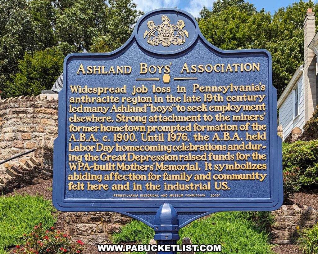 Historical marker for the Ashland Boys' Association, a blue and gold sign with the Pennsylvania Historical and Museum Commission emblem, detailing the association's history and its strong ties to the local community, with the WPA-built Mothers' Memorial mentioned. The marker is set against a landscaped background with a stone wall and residential buildings in Ashland, Pennsylvania.