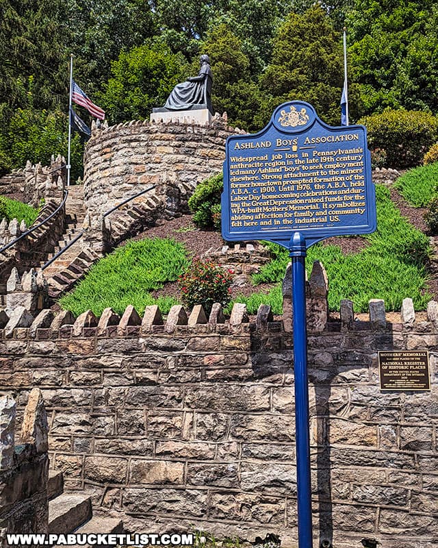 The Ashland Mothers' Memorial in Ashland, Pennsylvania, viewed from the base of the stone steps leading up to the statue. A historical blue and gold marker stands in the foreground, detailing the Ashland Boys' Association. Behind it, the bronze statue of a seated woman atop a circular stone wall is framed by an American flag on one side and green trees on the other. A plaque noting the memorial's placement on the National Register of Historic Places is also visible at the base of the signpost.