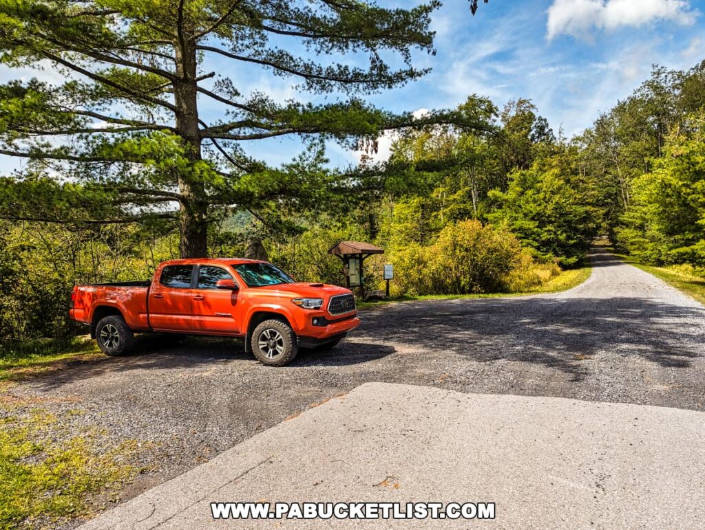 A vibrant orange pickup truck is parked on the side of a gravel road leading into the forested area of Bear Meadows Loop Hike in Centre County, Pennsylvania. The road forks ahead, surrounded by lush greenery and trees, under a partly cloudy sky. An information kiosk is partially visible on the left, suggesting a trailhead or parking area for hikers venturing into the natural landscape.