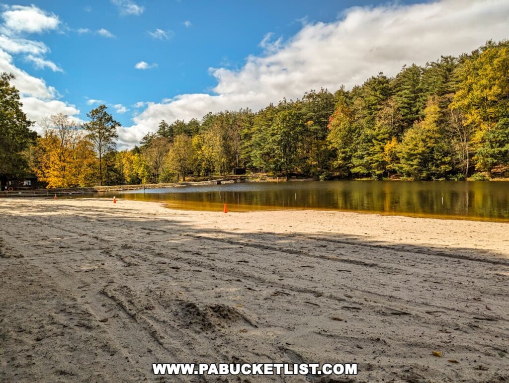 The sandy beachfront at Colonel Denning State Park in Cumberland County, PA, with tire tracks on the sand leading towards a tranquil body of water. The lake is bordered by trees with autumn leaves in shades of green, yellow, and orange, under a partly cloudy sky.