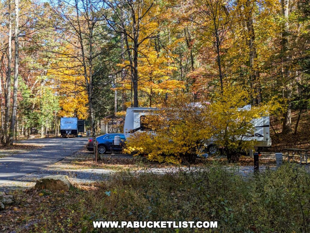 A campsite at Colonel Denning State Park in Cumberland County, PA, during autumn. The scene includes recreational vehicles parked among trees with colorful fall foliage in hues of yellow, orange, and red. The ground is covered with fallen leaves, and there's a crisp blue sky above.