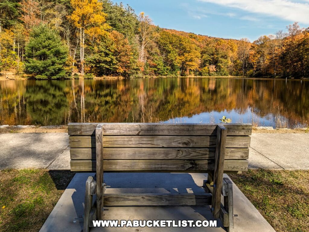 View from behind a wooden bench at Colonel Denning State Park in Cumberland County, PA, overlooking a peaceful lake reflecting the vivid colors of autumn trees. The foliage is a mix of green, yellow, and orange, creating a mirrored image on the water's calm surface on a bright sunny day.