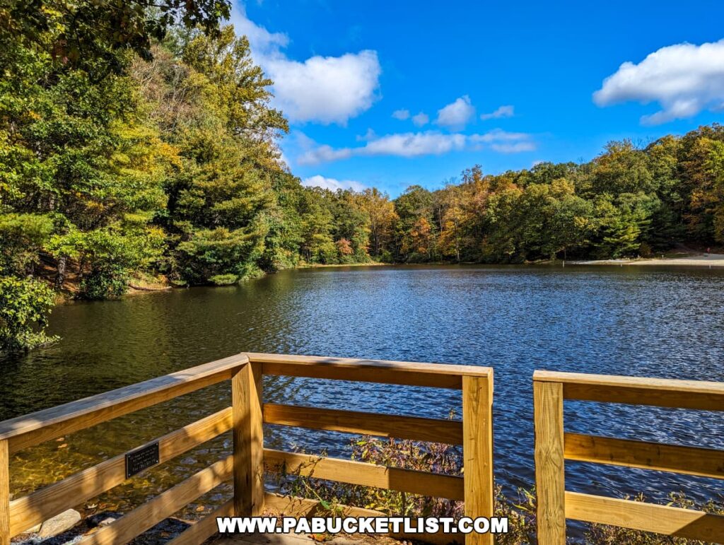 View from a wooden fishing pier at Colonel Denning State Park in Cumberland County, PA, overlooking a lake surrounded by dense forests with autumn foliage. The clear blue sky with fluffy clouds is reflected in the lake's surface, enhancing the peacefulness of this natural setting